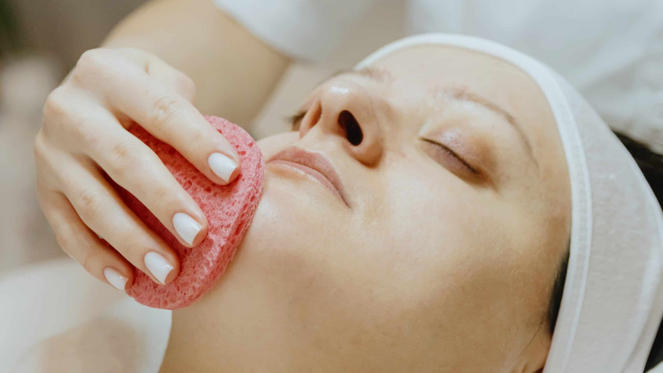 A young unrecognizable Caucasian girl cosmetologist cleans the chin of an adult female client's face using a pink scrubbing sponge, which lies on a massage table in a beauty salon, side view close-up.