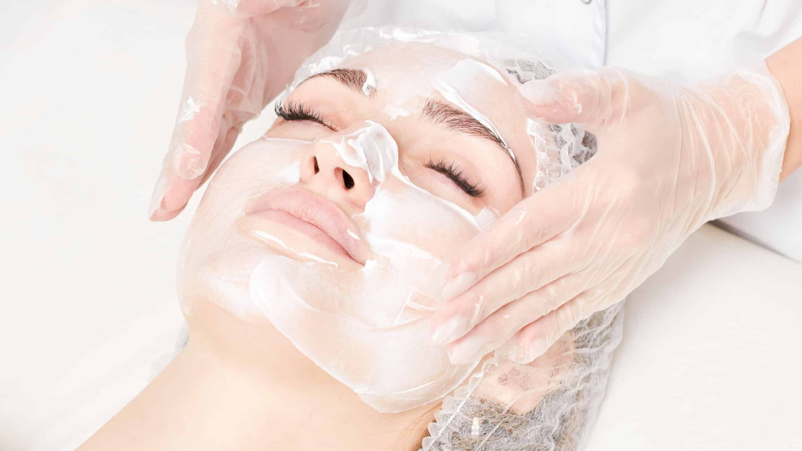 Beautician massages cream mask into female face skin for rejuvenation, moisturizing skincare cosmetic procedure in beauty spa salon. Cosmetologist hands in gloves rubbing cream in woman face